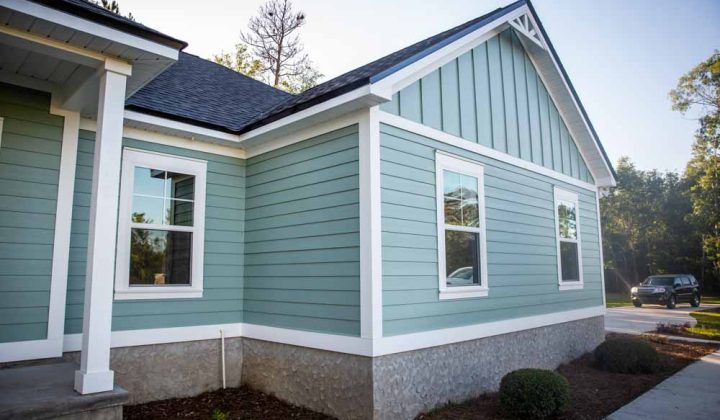 A view of a cottage style home with green siding, white trim, and a gable roof, surrounded by well-kept landscaping, presenting a quaint and welcoming exterior.